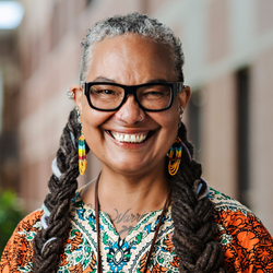 Headshot of Ann Marie Beals, a person wearing black-framed glasses smiling, wearing two thick braids, a colourful top and long colourful earrings.