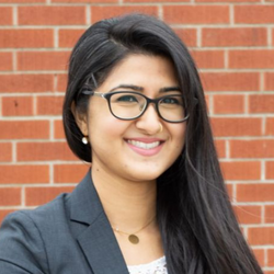 A headshot of Heena Mistry, a person smiling with long dark hair, dark-rimmed glasses, and a blazer.