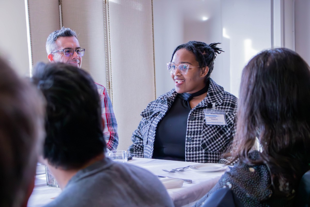 A Black graduate student talks to a table full of other professors and students.