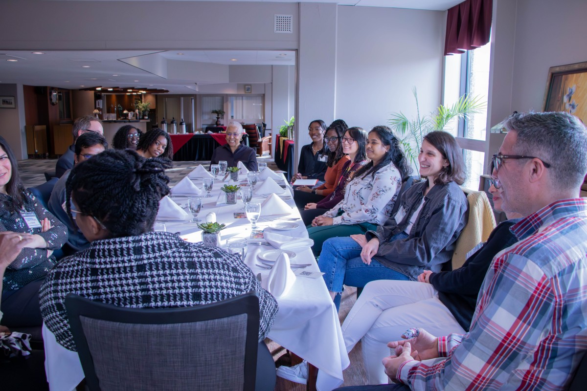 A group of diverse faculty and graduate students sit together at a long table set for dinner.