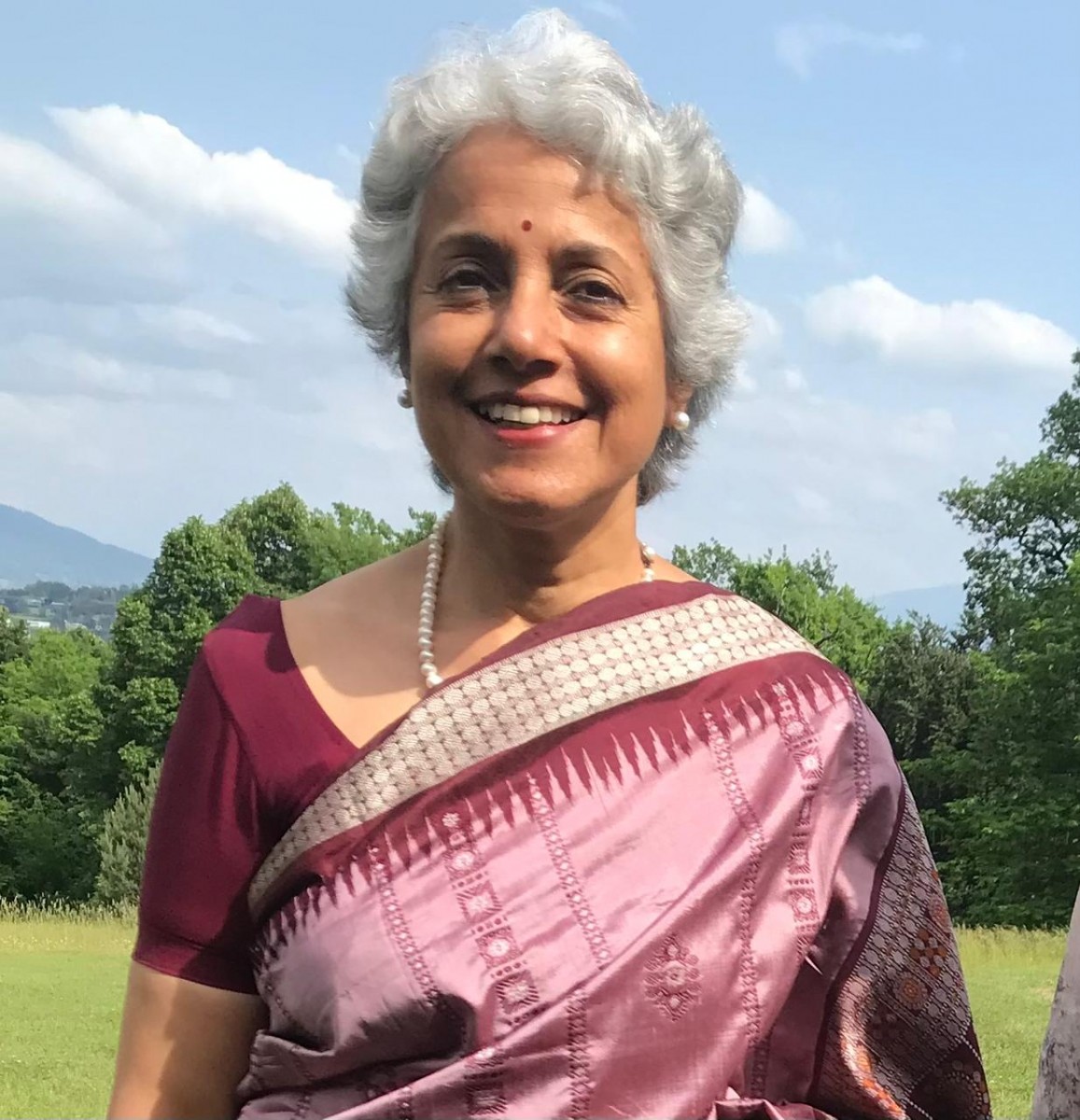 A headshot of Dr. Soumya Swaminathan, a medium-skinned woman with short, curly grey hair and wearing a pink sari smiling at the camera. She's standing outdoors in a field on a sunny day with trees in the distance.
