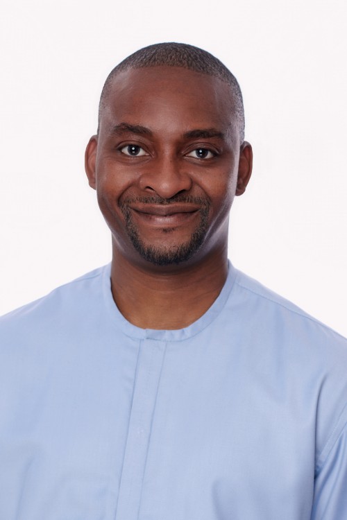 A headshot of Uwa Idemudia, a bald dark-skinned man who is smiling at the camera wearing a light-blue shirt.