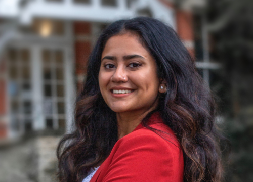 A headshot of Anindita (Dita), a South-Asian woman with long dark-brown and black hair wearing a red blazer, while being shifted to the side smiling into the camera. 
