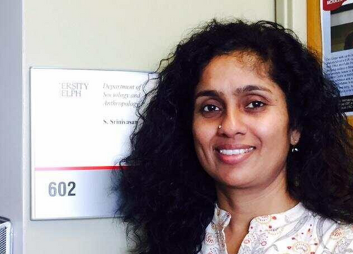 A headshot of Sharada Srinivasan, a woman with light brown skin and thick black curly hair smiling at the camera