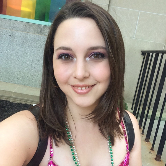 A photo of Abigail dressed for Pride in a sequin tank top and mardi gras beads in front of a pride flag.