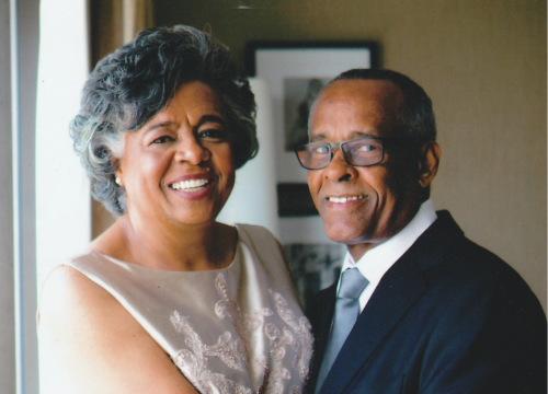 The Brathwaites stand by a window dressed in formal attire and in a sideways embrace while smiling at the camera.