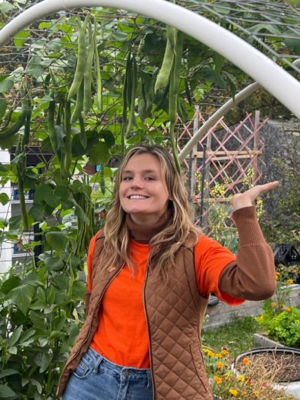 Dakota Cherry poses cheekily for a photo in front of an archway of plants, wearing an orange shirt and a brown vest and smiling.