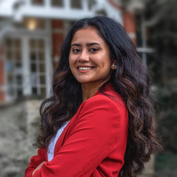 A headshot of Anindita (Dita), a South-Asian woman with long dark-brown and black hair wearing a red blazer, while being shifted to the side smiling into the camera.