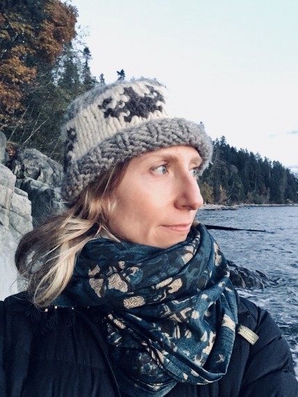 Justine Townsend in toque and scarf stands on a beach looking out at the water