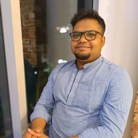 M Manjurul Islam sits at a table and smiles at the camera. He has medium-brown skin, short black hair, a goatee, glasses, and a blue long sleeve shirt.