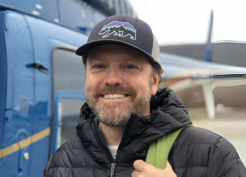 Nicholas stands in front of a helicopter wearing a puffer coat, a baseball cap and a backpack, smiling at the camera.