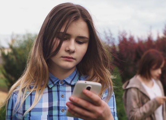 sad young girl checking messages on her phone. Other young girl in the background.